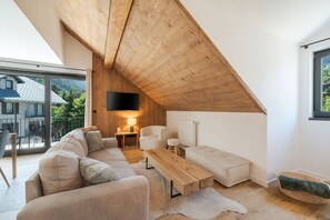 A modern loft bedroom with a sloped ceiling, a large window, a flat-screen TV, a sofa, a coffee table, and a view of a balcony with outdoor furniture.