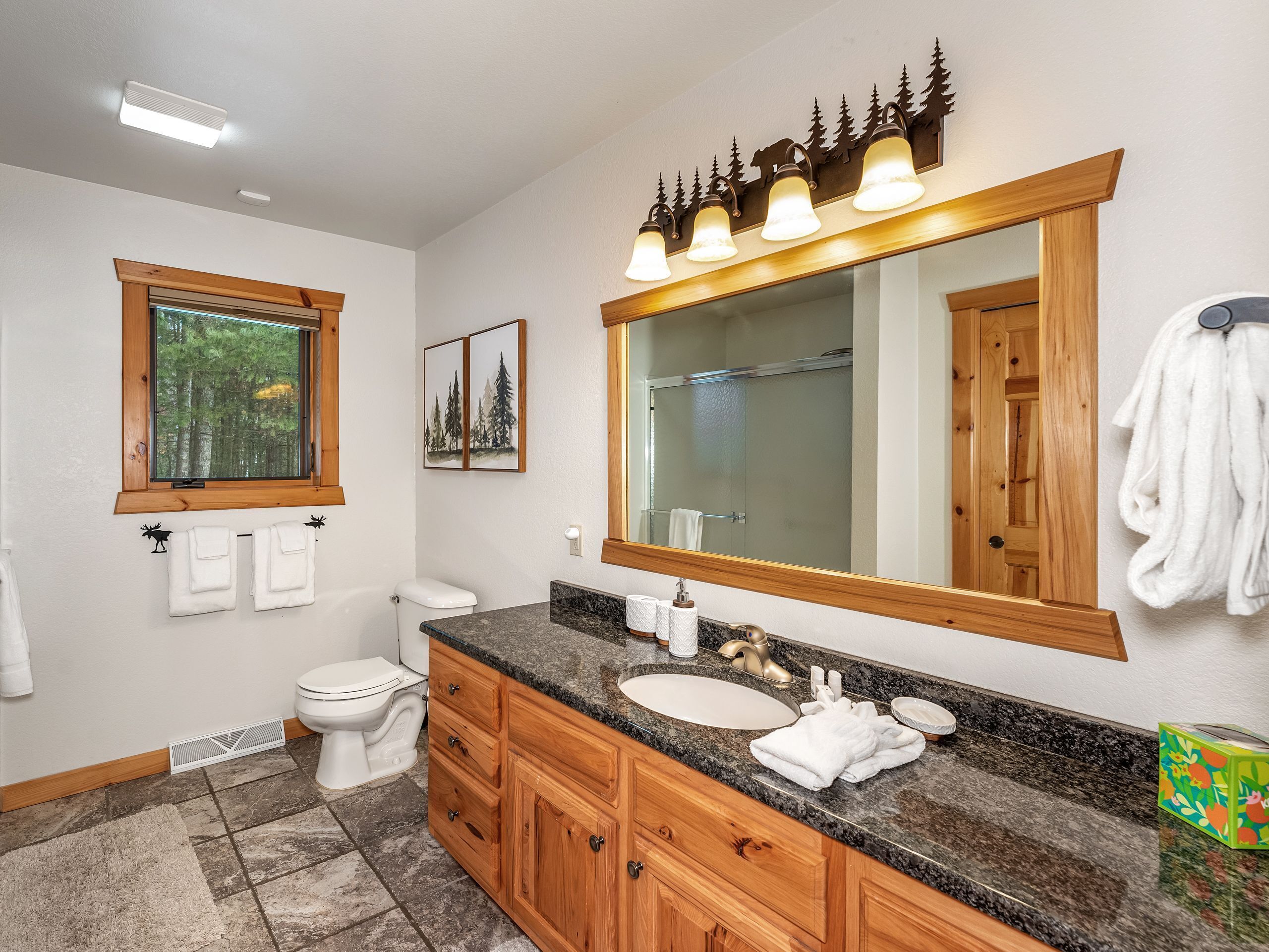 A bathroom with a wooden vanity, a large mirror, a toilet, and a window with a view of trees.