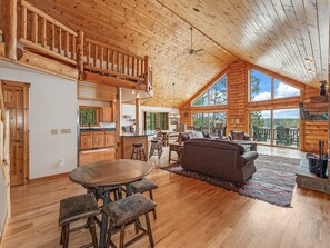 Una cabaña de troncos espaciosa con techo alto, pisos de madera, zona de comedor con mesa redonda y taburetes, sala de estar con sofá y sillones, y ventanas amplias con vistas al bosque circundante.