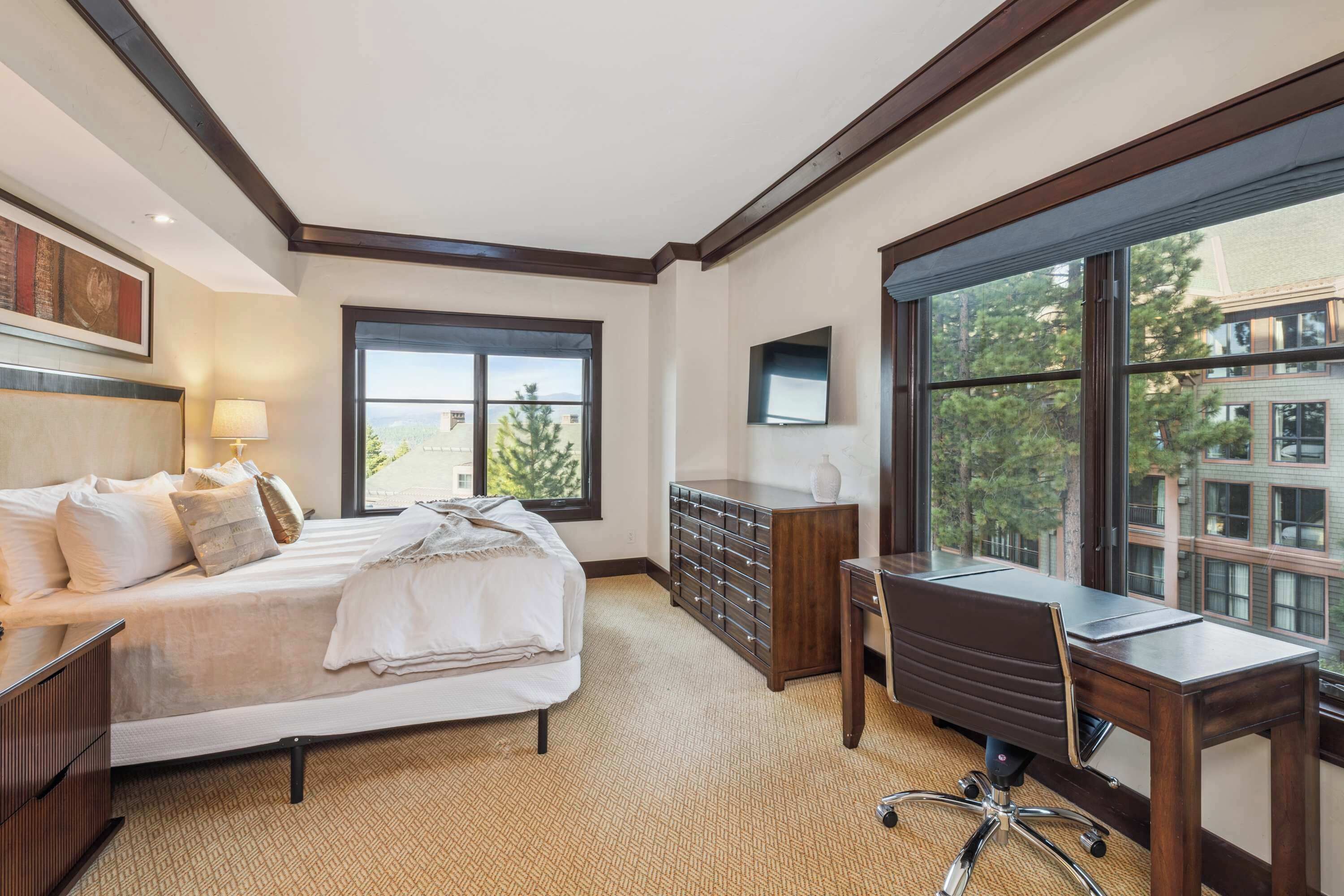 A hotel room with a bed, desk, dresser, and large windows offering a city view.