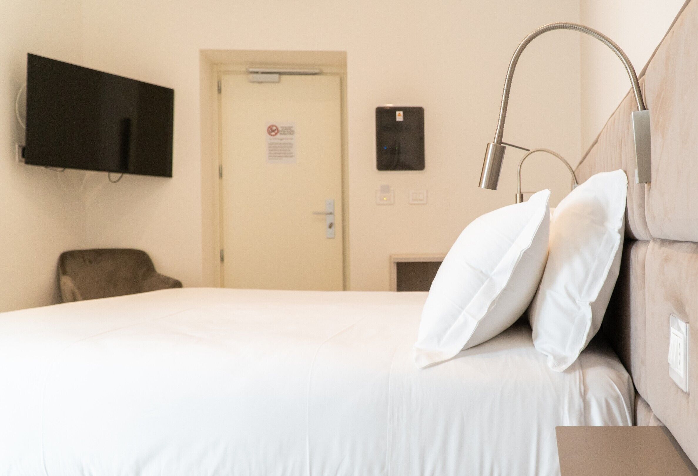 A hotel room with a bed, a chair, a television, a door, and a control panel.
