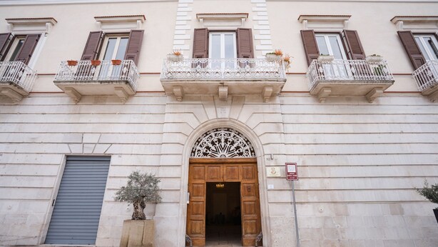 Une façade d’immeuble avec une entrée centrale, des balcons dotés de garde-corps et un pot de fleurs.
