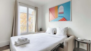 A hotel room with a bed, a nightstand, a lamp, a window with curtains, and a painting on the wall. - Cosy Studio Asnieres (Asnières-sur-Seine)
