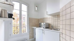 A compact kitchen with white cabinets, a built-in microwave, and a window with a view of buildings. - Cosy Studio Asnieres (Asnières-sur-Seine)