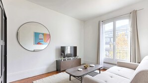 A modern living room with a sofa, a coffee table, a TV on a cabinet, a round mirror, and a window with curtains. - Cosy Studio Asnieres (Asnières-sur-Seine)