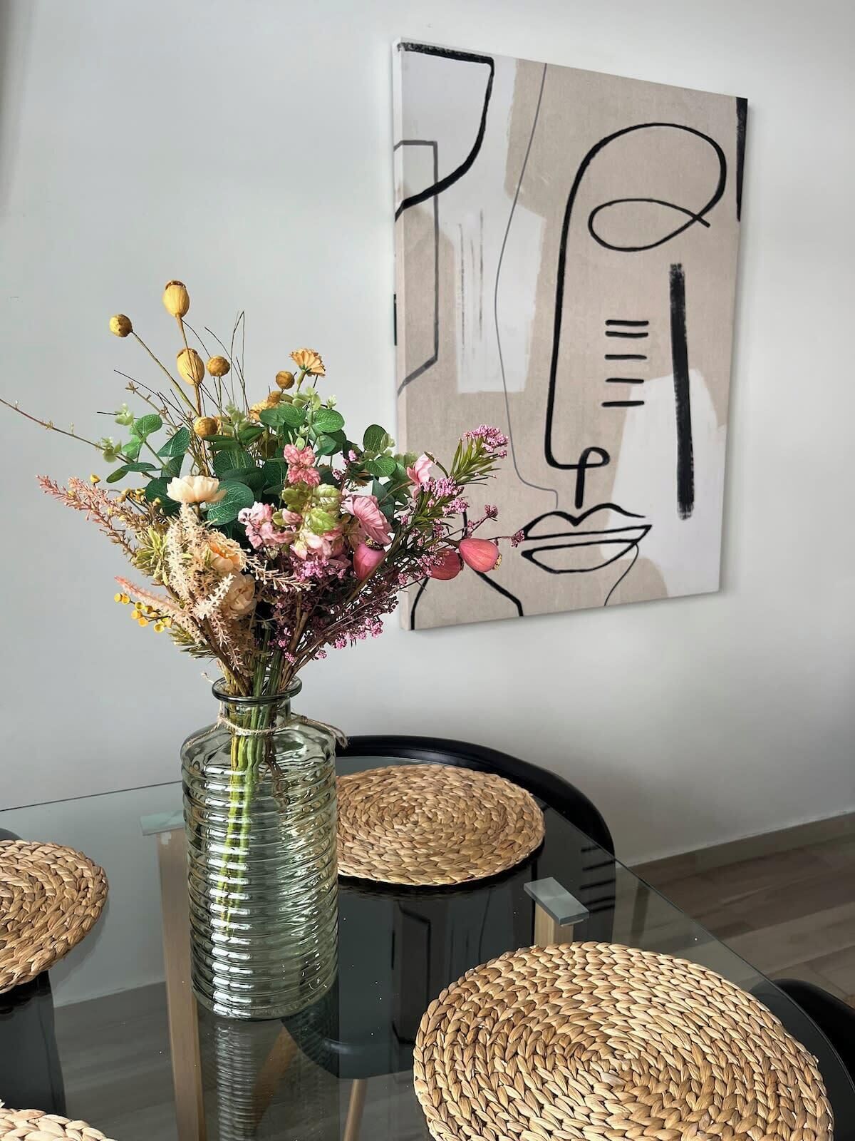 Una mesa de comedor con un florero de cristal, individuales tejidos y una pared con arte abstracto.