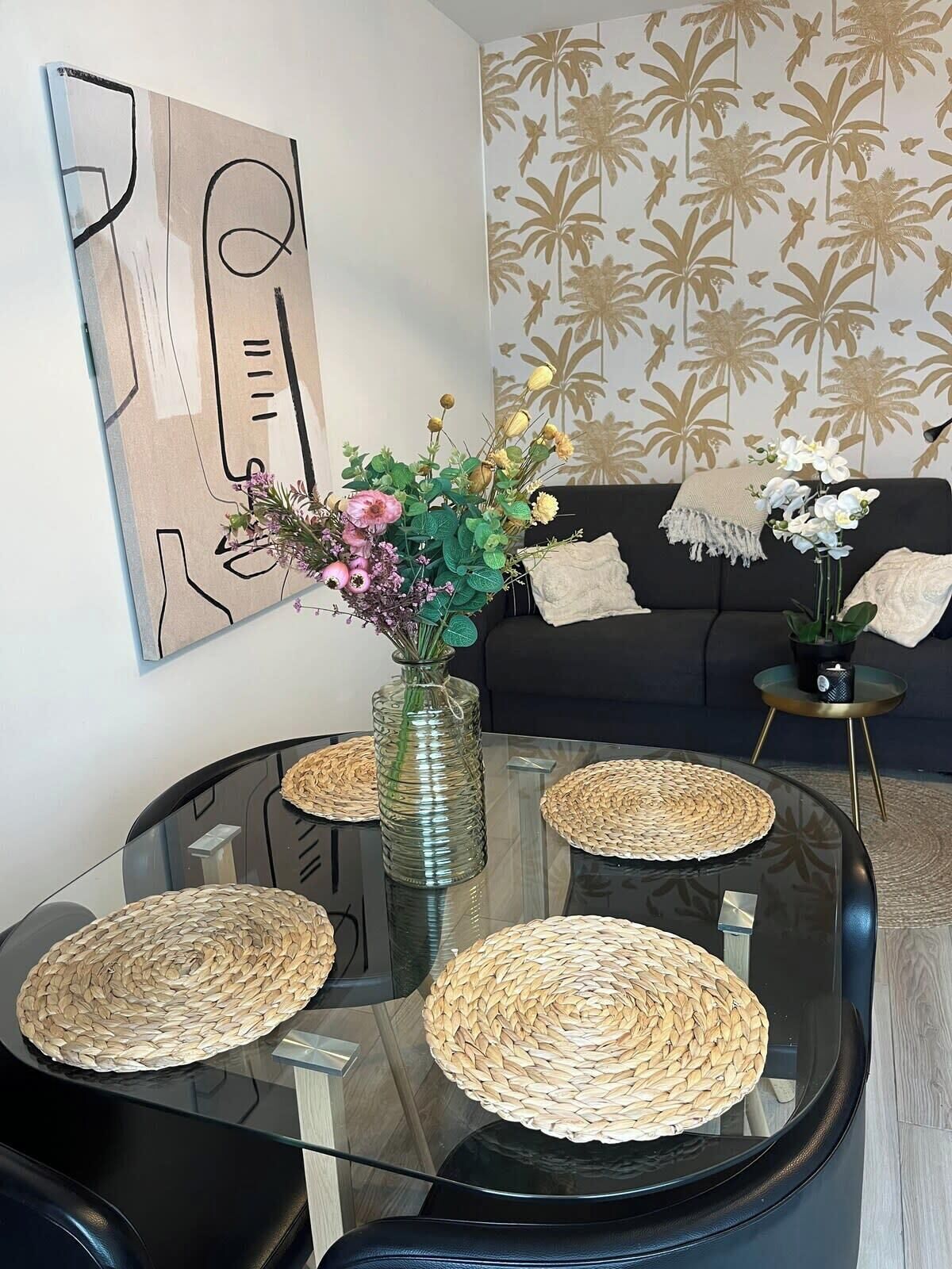 Un salon moderne avec une table basse en verre, des sets de table en fibres tressées, un vase avec des fleurs et un papier peint à motifs.