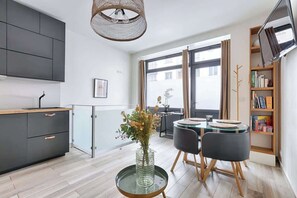 Eine moderne Küche mit dunklen Schränken, einem Glastisch mit Stühlen, einer Vase mit Blumen und einem Bücherregal.