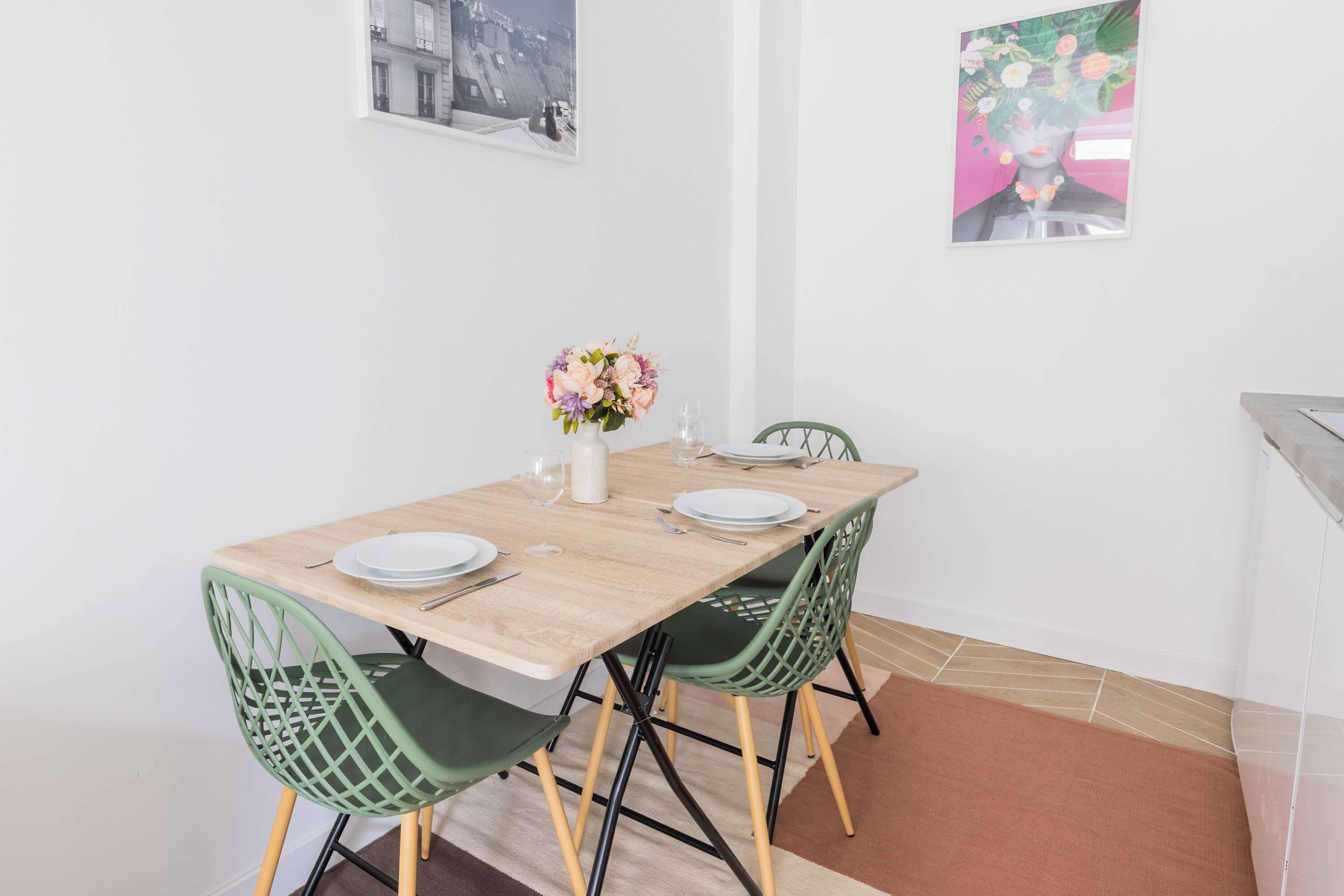 A dining area with a wooden table set for two, green chairs, a vase with flowers, and two framed pictures on the wall.
