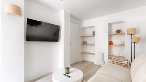 A modern living room with a flat-screen TV mounted on the wall, a round coffee table with a remote control, a beige sofa, and built-in shelves with decorative items. - Modern Studio Near Paris w/ Fast WiFi, TV (Charenton-le-Pont)