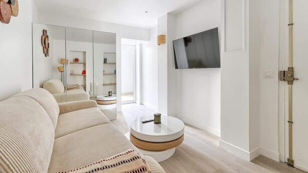 A modern living room with a sofa, a round coffee table, a flat-screen TV mounted on the wall, and a door with a handle. - Modern Studio Near Paris w/ Fast WiFi, TV (Charenton-le-Pont)