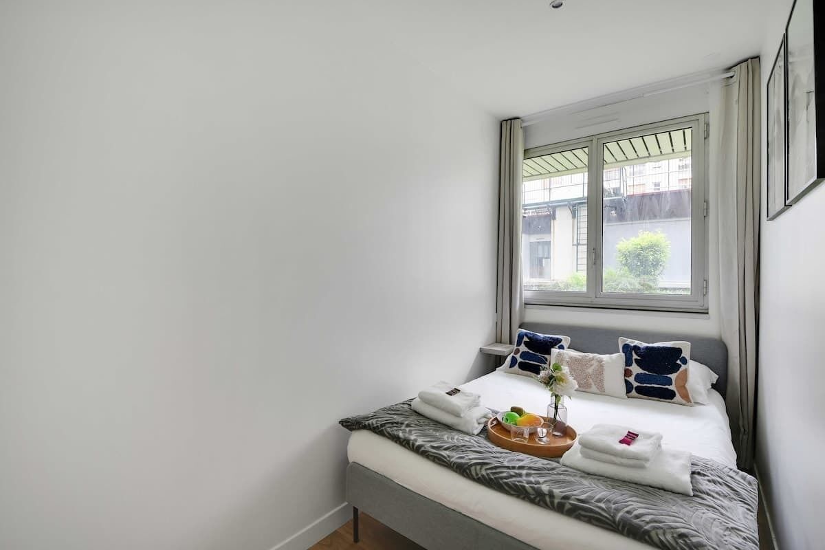 A neatly made bed with a tray of fruits and a glass of water, a window with a view of buildings, and a framed picture on the wall.