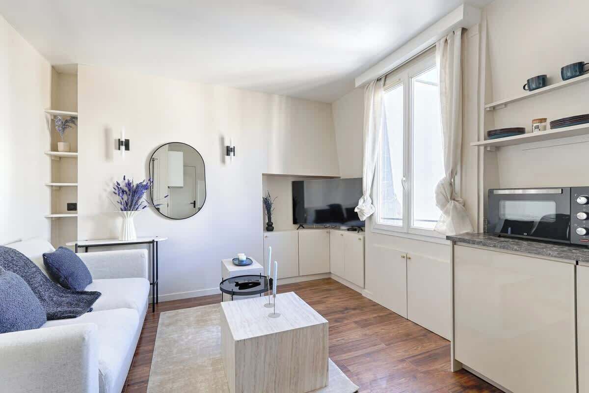 A modern living room with a sofa, coffee table, and a TV. There is a kitchen area with cabinets and a microwave.