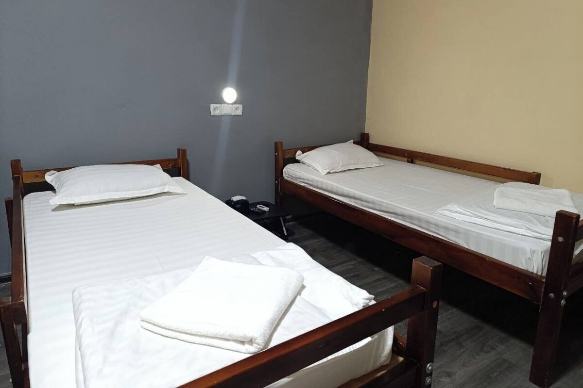 A room with two wooden beds, each with white linens and pillows, a small bedside table, and a wall-mounted light fixture.