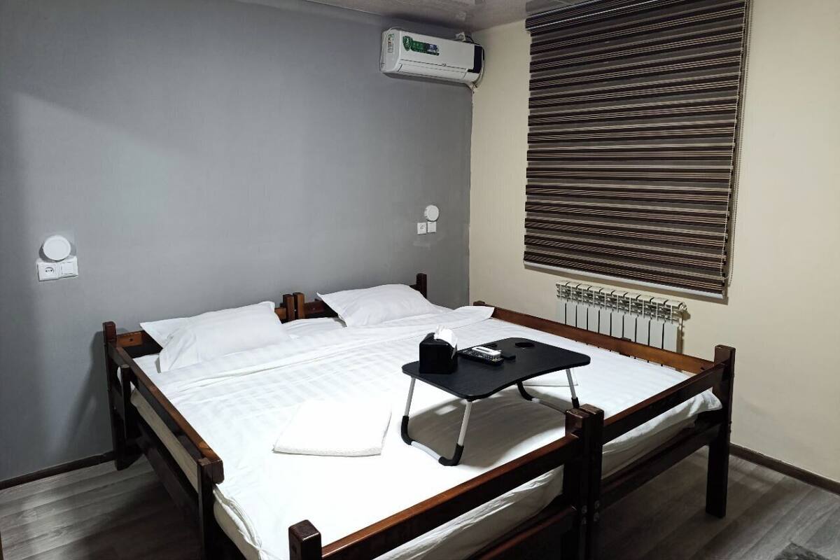 A single bed with a wooden frame, a bedside table with a remote control, a pillow, and a blanket. There is a window with blinds, a radiator, and an air conditioning unit mounted on the wall.