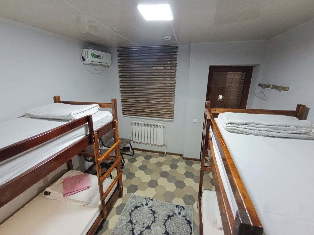 A dormitory room with bunk beds, a radiator, a window with blinds, and a hanging coat rack.