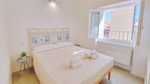 A neatly made bed with rolled towels, a small bedside table with a plant, and a window with a view of the outside.