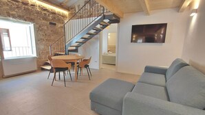 A living room with a grey sofa, a dining table with chairs, a staircase, and a TV mounted on the wall.
