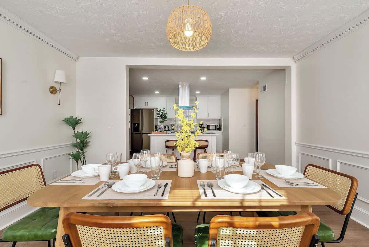 A dining area with a wooden table set for four, surrounded by wicker chairs, and a hanging light fixture above.