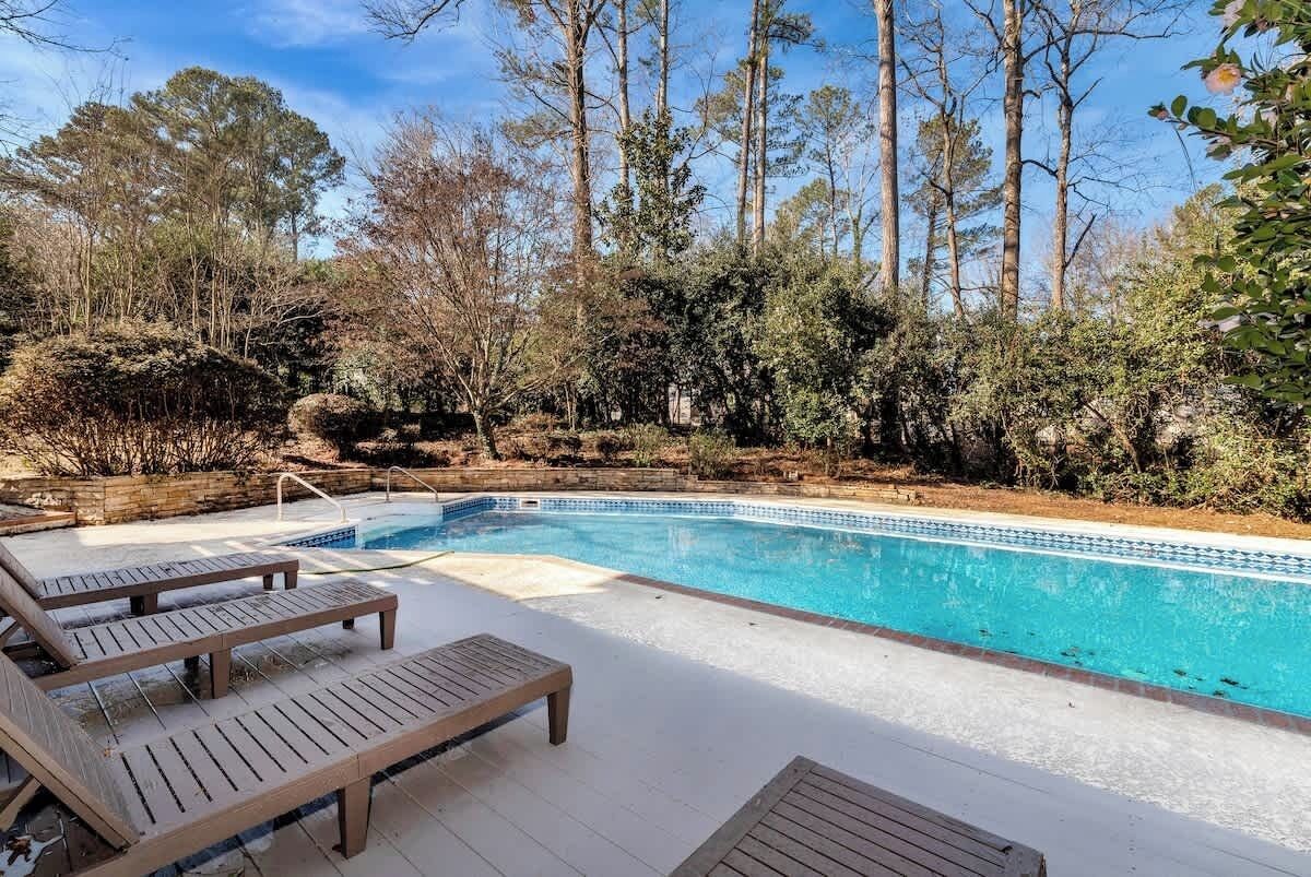 Une piscine entourée d’arbres et d’arbustes, avec des chaises longues en bois disposées tout autour.