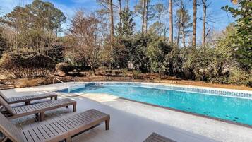 Une piscine entourée d’arbres et d’arbustes, avec des chaises longues en bois disposées tout autour.