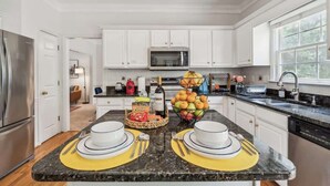 A modern kitchen with white cabinets, a stainless steel refrigerator, a microwave, and a sink. There are two dining areas with yellow placemats and white plates. - Chic Metro Family Stay / High Ceiling / Game Room (Lawrenceville)
