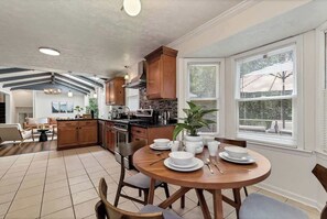Une cuisine moderne avec des meubles en bois, une table à manger pour quatre personnes et une grande fenêtre équipée de stores.