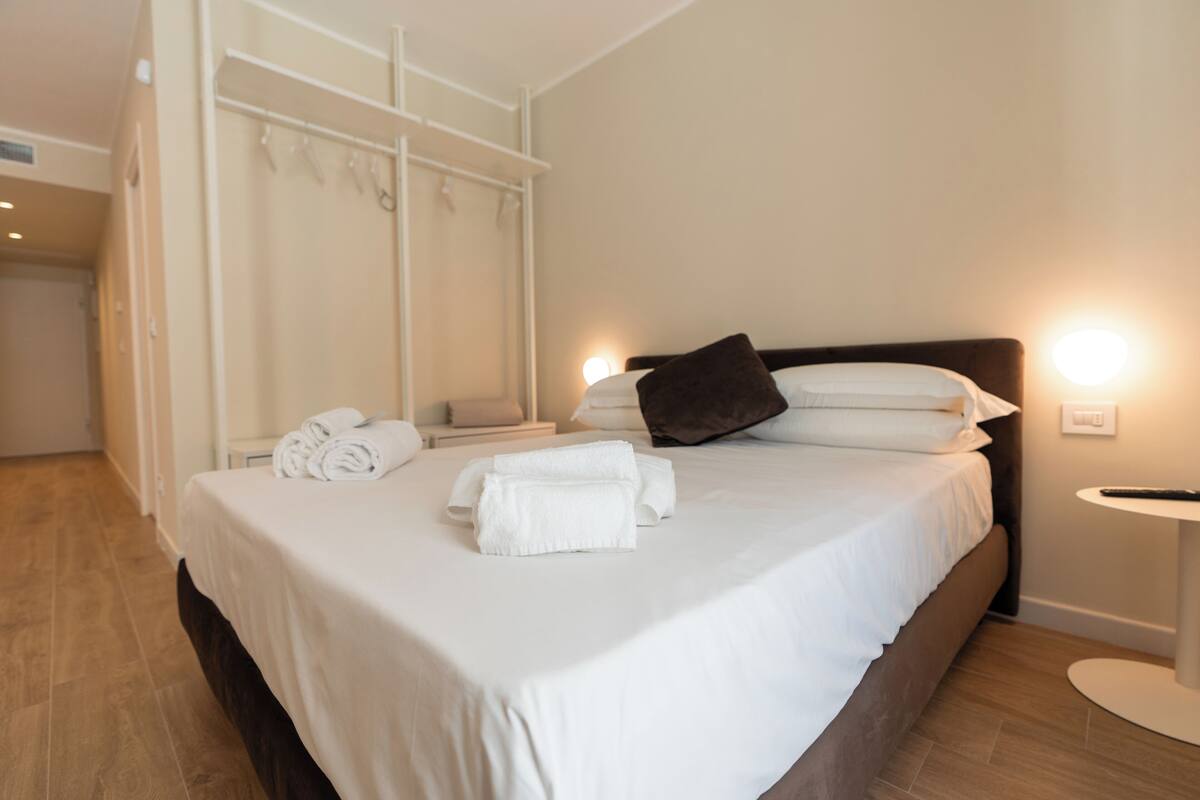 A neatly made bed with white linens and towels, a bedside table with a remote control, and a wall-mounted coat rack.