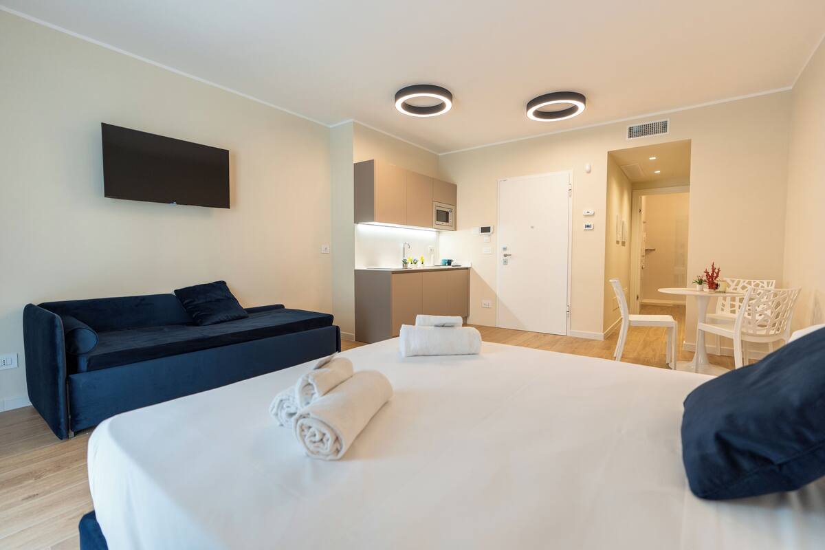 A modern hotel room with a bed, a sofa, a TV, and a kitchenette.