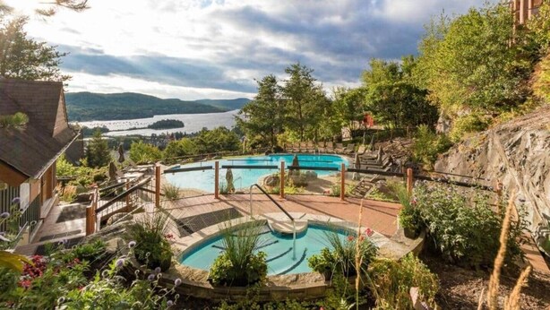 A pool area with a view of a lake and mountains, surrounded by greenery and a wooden deck. - Equinoxe 152-3 Ski in/Ski out (Mont-Tremblant)