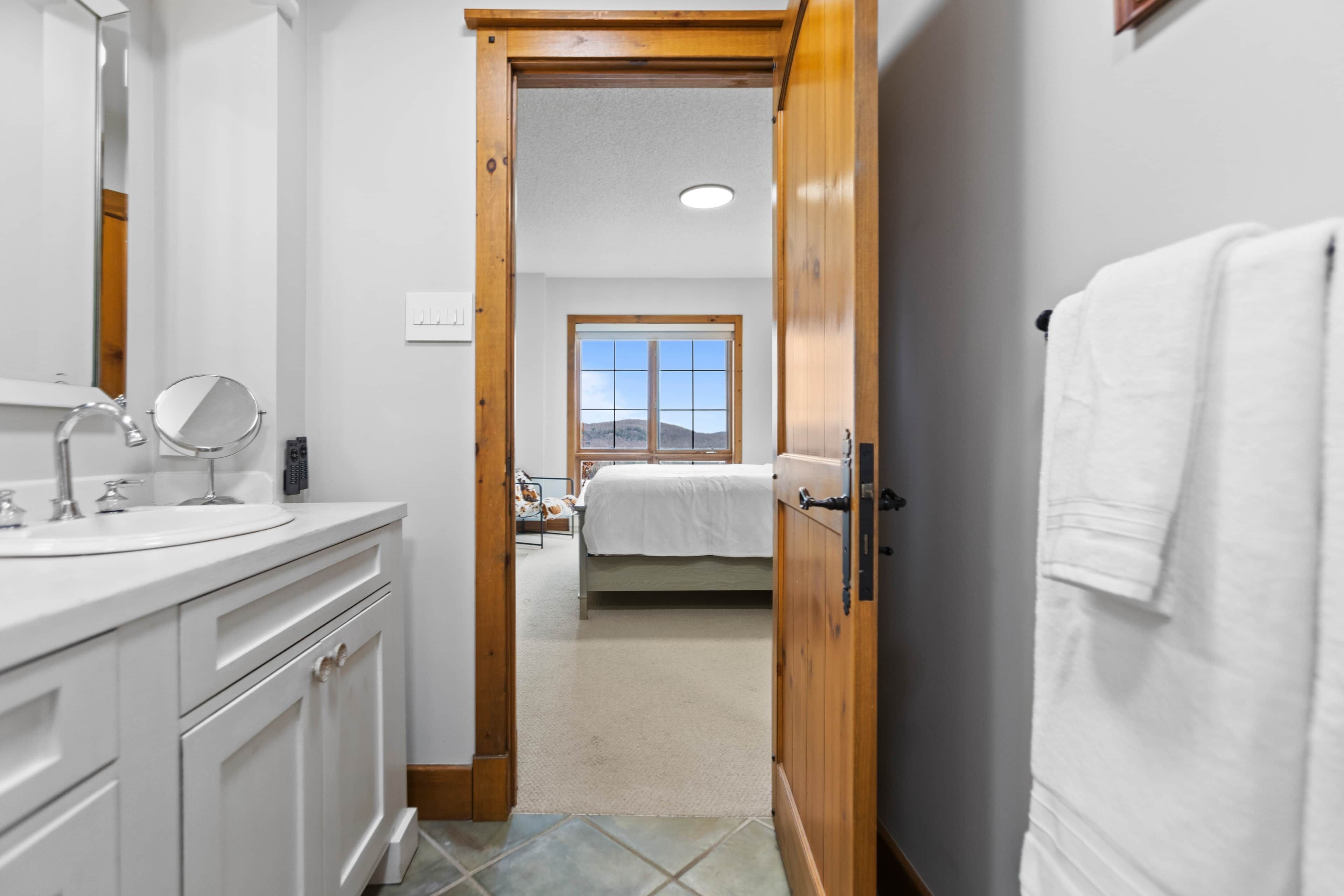 Une salle de bain avec un lavabo blanc, un miroir et une porte en bois donnant sur une chambre à coucher équipée d’un lit et d’une fenêtre.