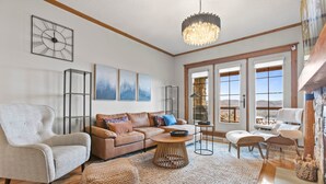 A spacious living room with a large sofa, a coffee table, and a fireplace. There are large windows and a chandelier. - Equinoxe 152-3 Ski in/Ski out (Mont-Tremblant)
