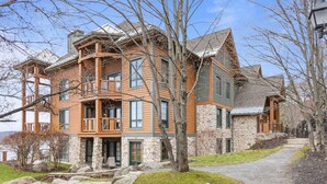 A large, multi-story house with a stone facade and wooden accents, featuring a balcony and a stone pathway leading to the entrance. - Equinoxe 152-3 Ski in/Ski out (Mont-Tremblant)