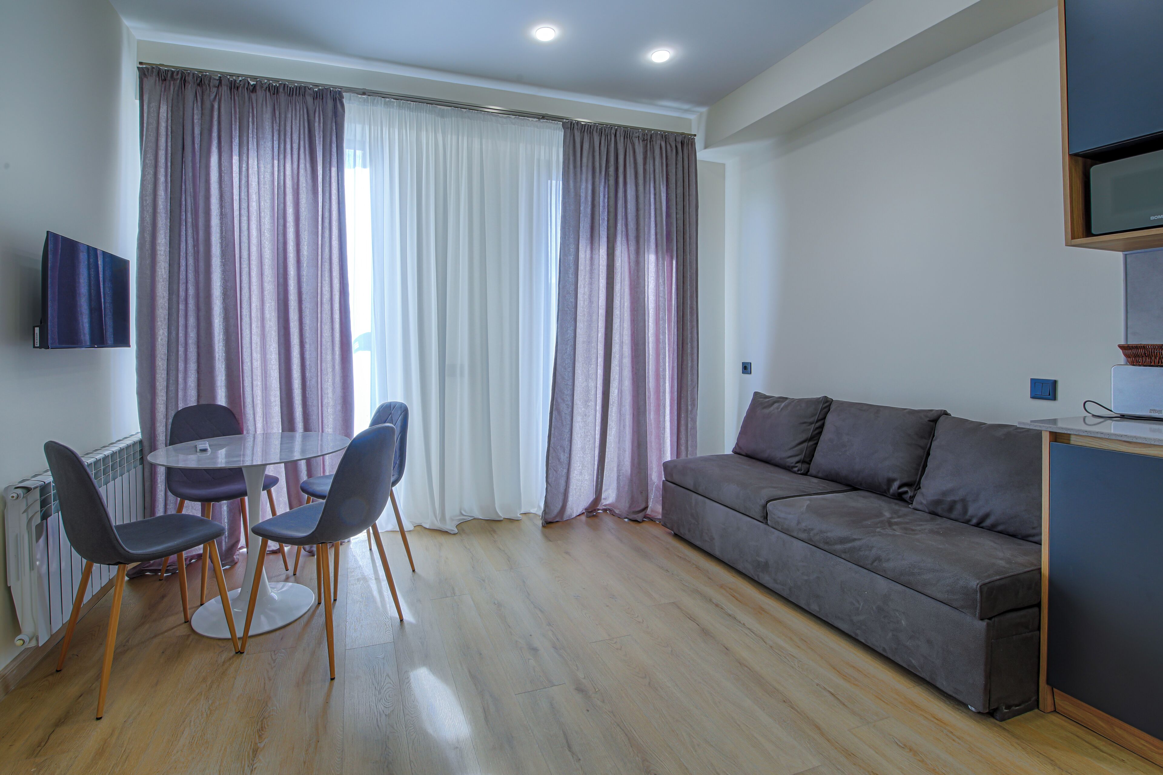 A modern living room with a grey sofa, a round dining table with four chairs, and a TV mounted on the wall.