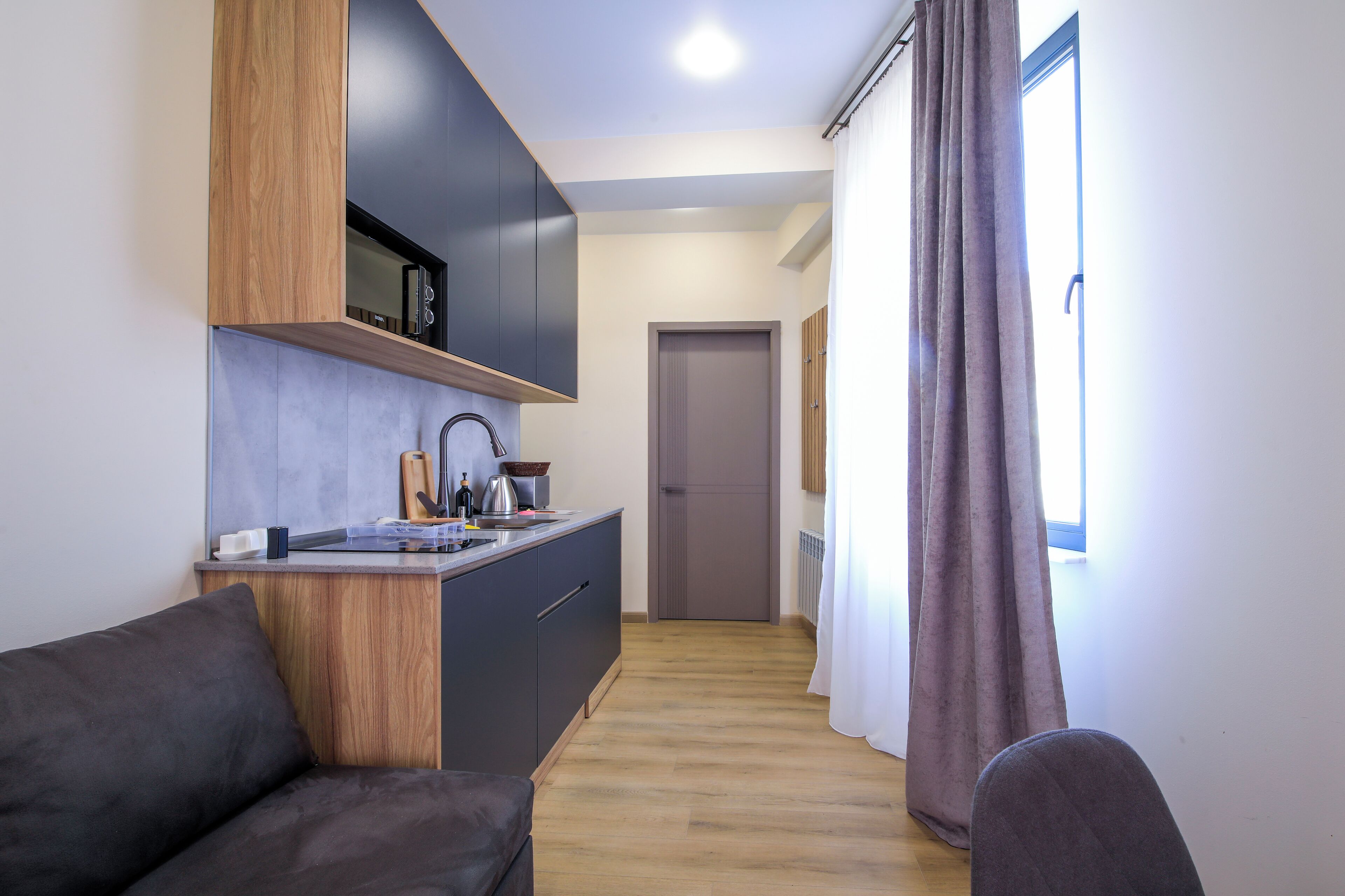 Una cocina moderna con microondas, pileta y muebles de madera. Un sofá oscuro está en primer plano, y se ve una ventana con cortinas.