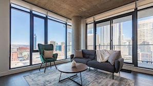 Un salón moderno con ventanas amplias, un sillón verde, un sofá oscuro y una mesa de centro redonda.