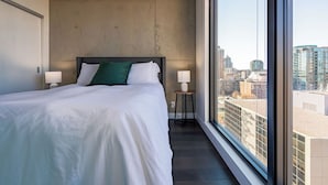 Habitación de hotel moderna con una cama grande, vista de la ciudad a través de un ventanal amplio y una mesita de noche con lámpara.