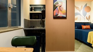 Cocina compacta con microondas, tetera y lavabo. Hay una silla verde y una mesa de madera. Se ve un sofá con un cojín 'OMAR' al fondo.