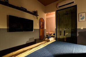 A hotel room with a bed, a flat-screen TV mounted on the wall, a small table with a chair, and a shower area. - Mini Studio 50m From the Beach AS 127/202b (Rio de Janeiro)