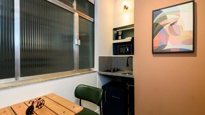 Cocina compacta con refrigerador, microondas y estufa. Hay una ventana con persianas, una mesa pequeña con una bicicleta y una silla.