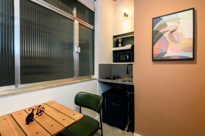 A compact kitchen with a refrigerator, microwave, and stove. There's a window with blinds, a small table with a bicycle, and a chair. - Mini Studio 50m From the Beach AS 127/202b (Rio de Janeiro)