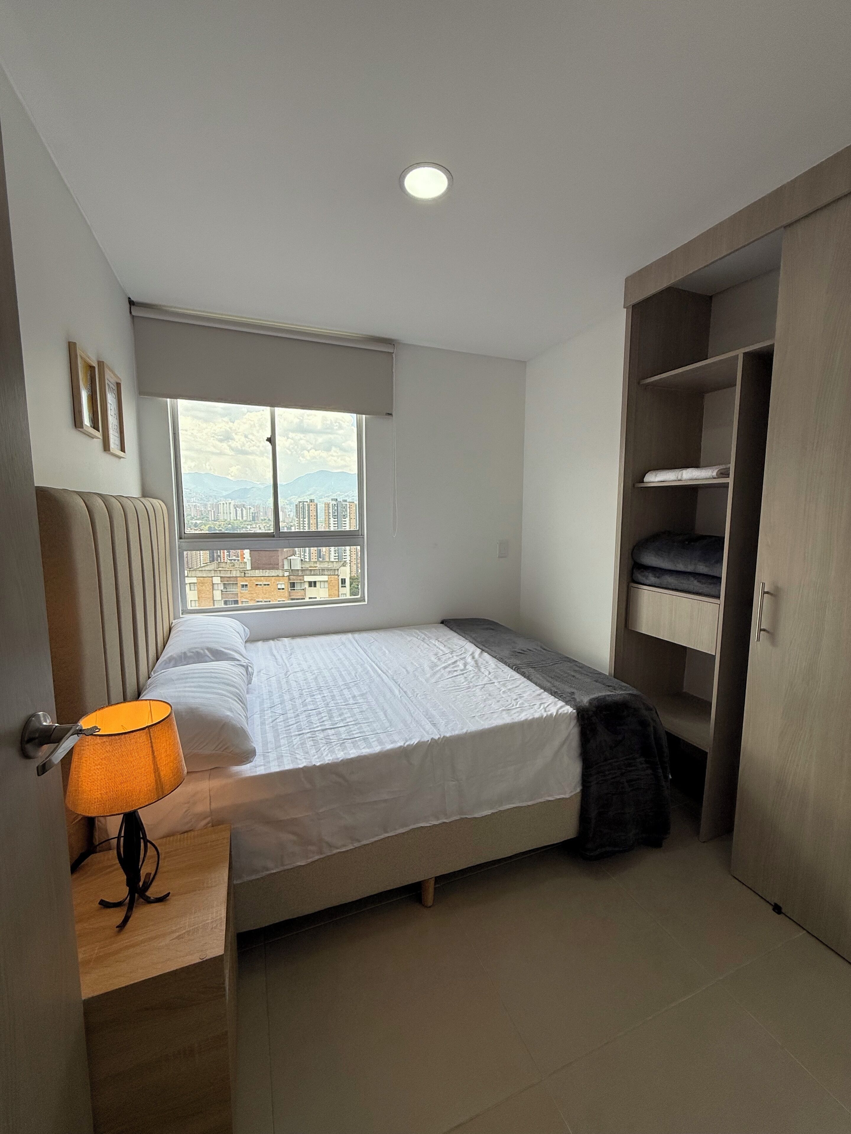 A bedroom with a bed, a bedside lamp, a window with a city view, and a wardrobe.