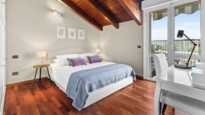 Een slaapkamer met een houten plafond, een bed met een blauwe deken, een bureau met een lamp en een raam met uitzicht op gebouwen.