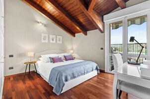 A bedroom with a wooden ceiling, a bed with a blue blanket, a desk with a lamp, and a window with a view of buildings.