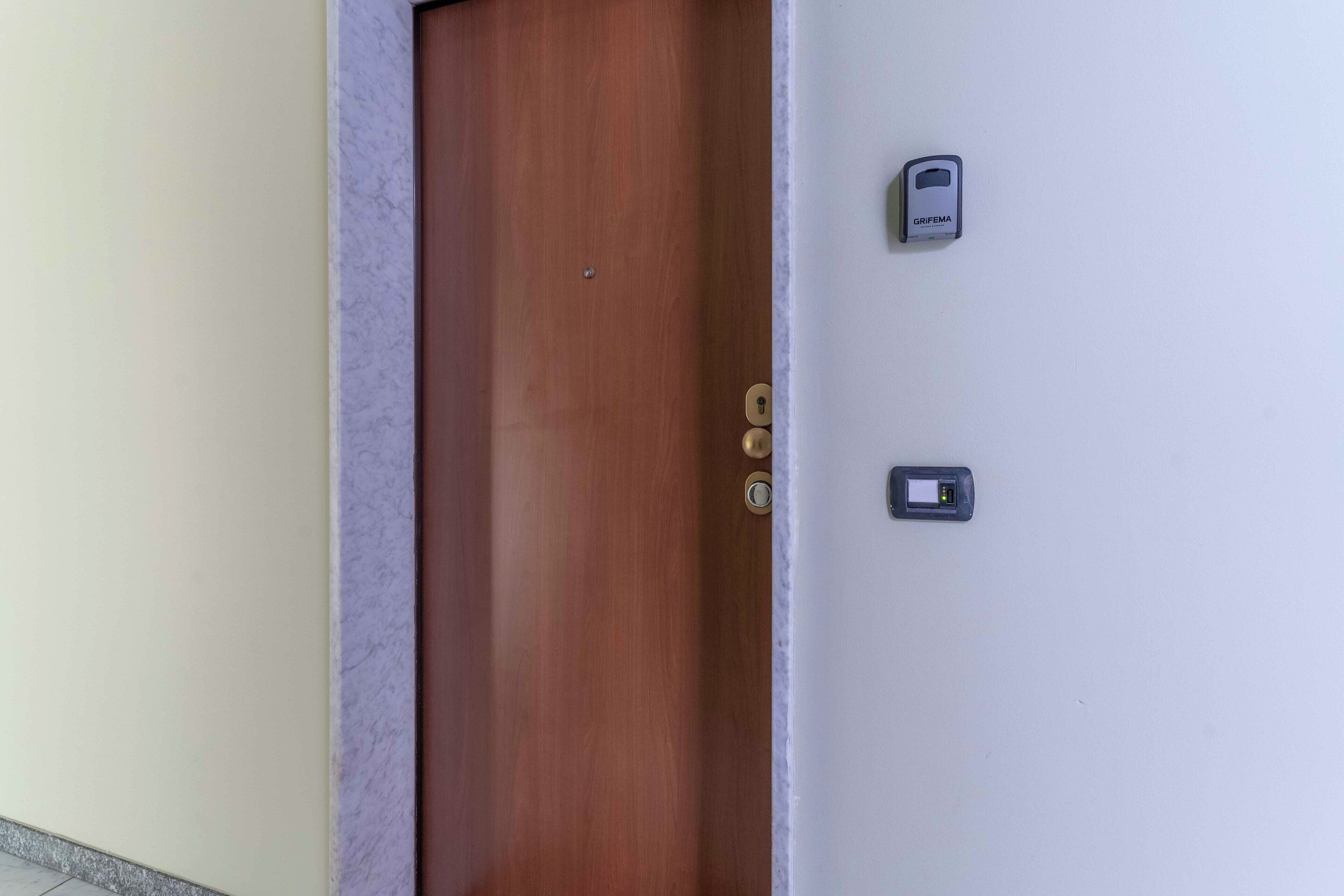 Una puerta de madera con manija de latón y cerradura, una pared blanca con un dispositivo GRIFEMA, y un objeto rectangular pequeño en la pared.