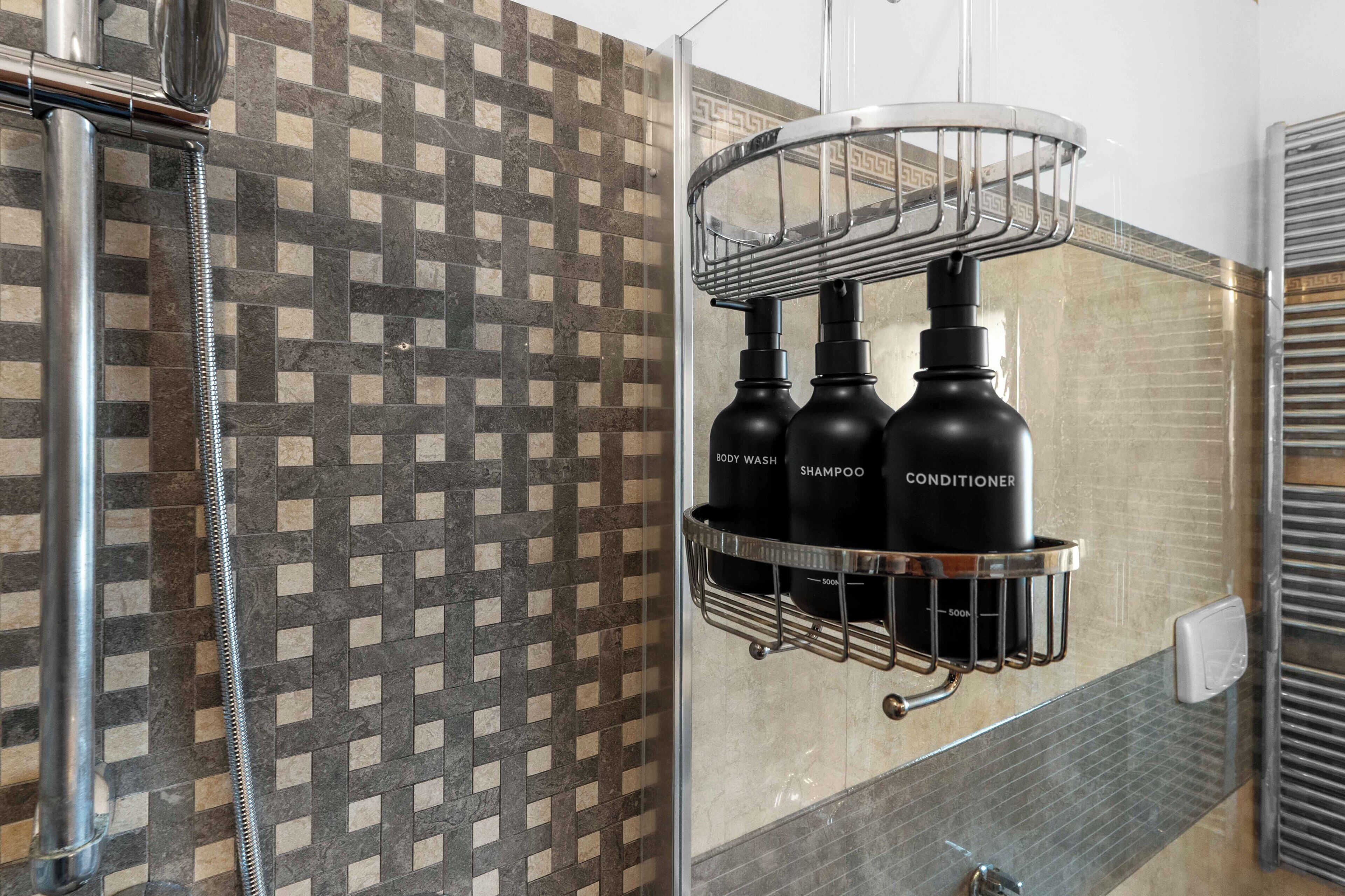 Un baño con pared de azulejos estampados, zona de ducha y estante con botellas de gel de baño, champú y acondicionador.