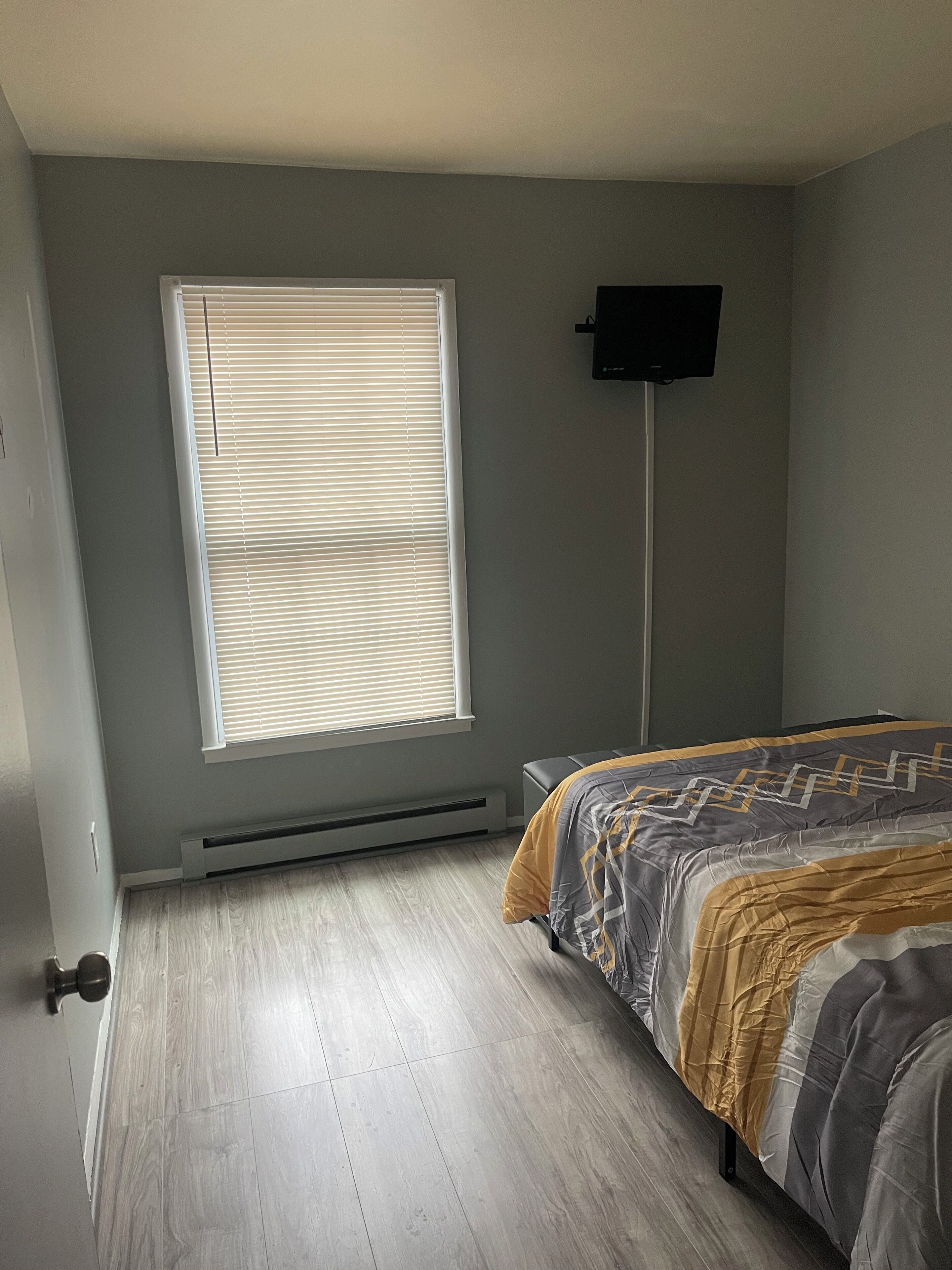 Une chambre à coucher avec un lit, une fenêtre avec des stores, un téléviseur fixé au mur et un radiateur.