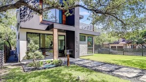 Een modern herenhuis met twee verdiepingen, een glazen voordeur, een stenen pad dat naar de deur leidt, en een kleine tuin met een boom en struiken.