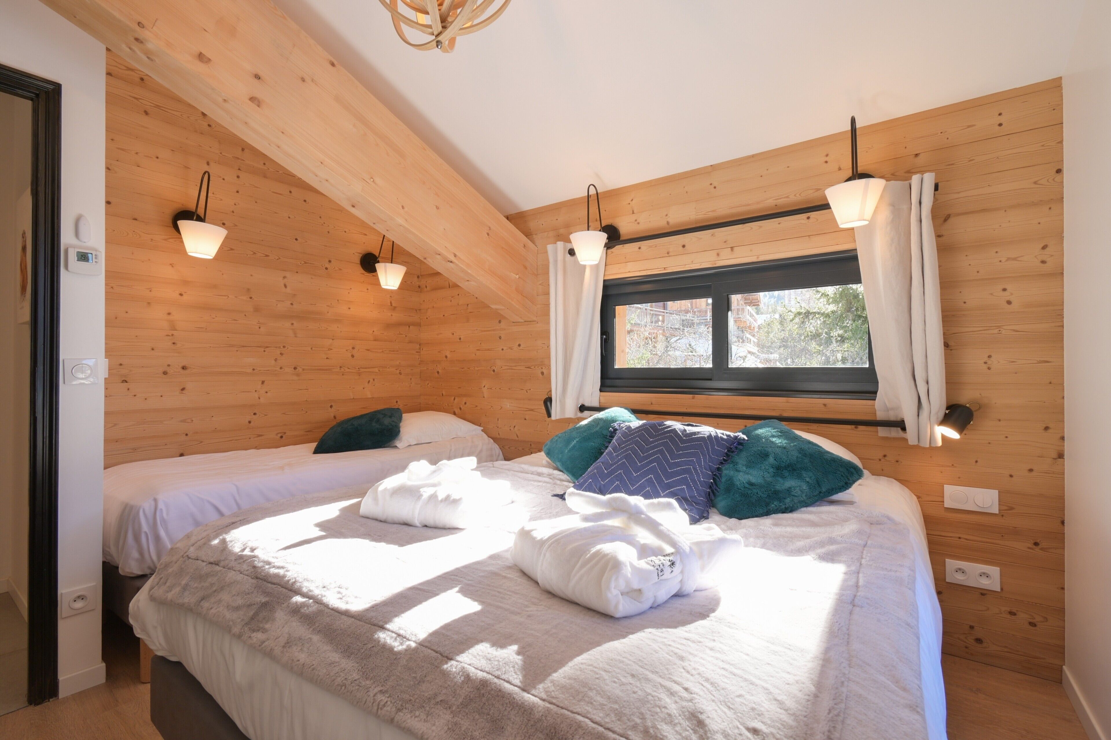 A room with wooden walls, two beds, a window with curtains, and wall-mounted lights.