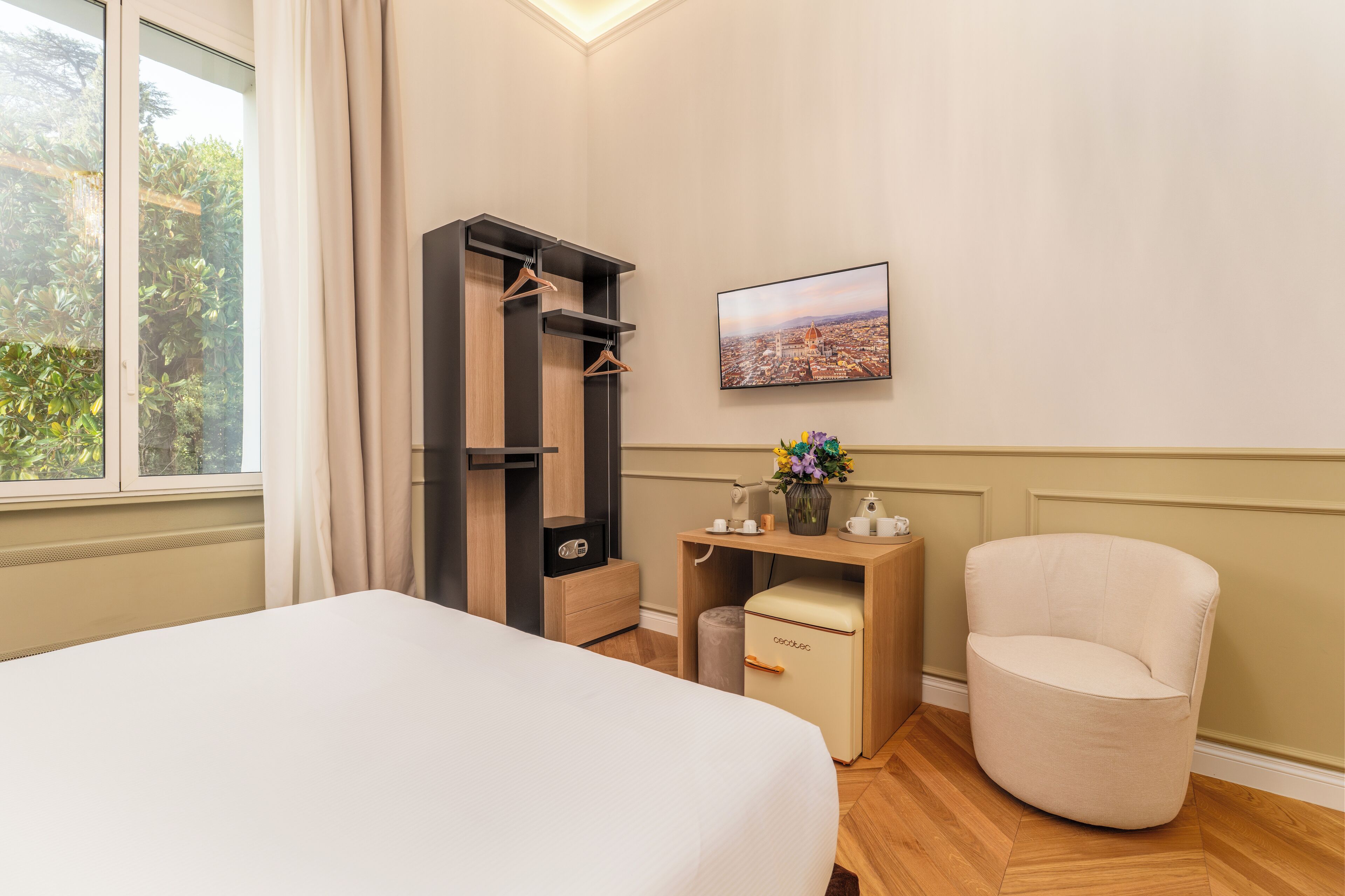 Habitación de hotel con cama, escritorio con silla, armario, una mesita con un florero y un cuadro de paisaje urbano en la pared.